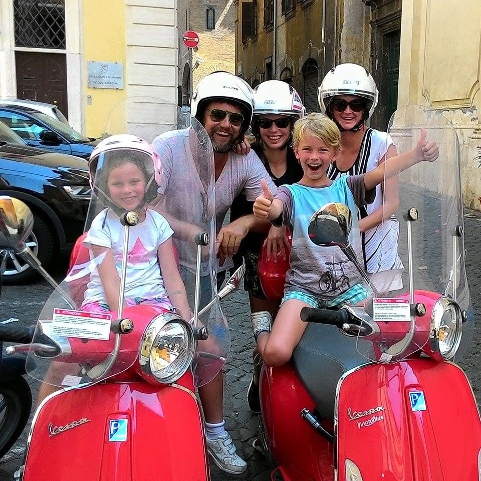 A family with two children in helmets, beside red Vespas in a Rome street