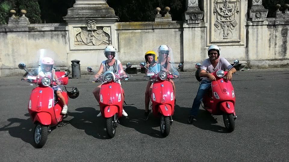 Four red Vespas lined up, children in helmets with their parents